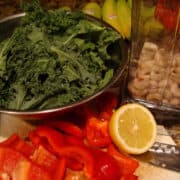 Fresh kale, chopped red bell pepper, sliced lemon, and a container of mushrooms on a kitchen countertop.