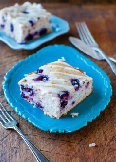 A piece of blueberry cake with icing on a blue plate, with forks and another slice in the background.