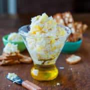 A glass of shredded cheese with a cork stopper on a wooden surface, surrounded by crackers and a small green bowl.