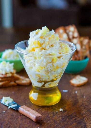 A glass of shredded cheese with a cork stopper on a wooden surface, surrounded by crackers and a small green bowl.
