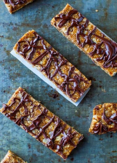 Seven homemade granola bars with a chocolate drizzle displayed on a blue surface.