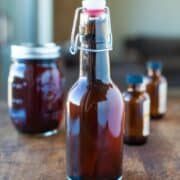 A small glass bottle with a swing-top closure filled with amber liquid, accompanied by smaller bottles and a jar in the background on a wooden surface.