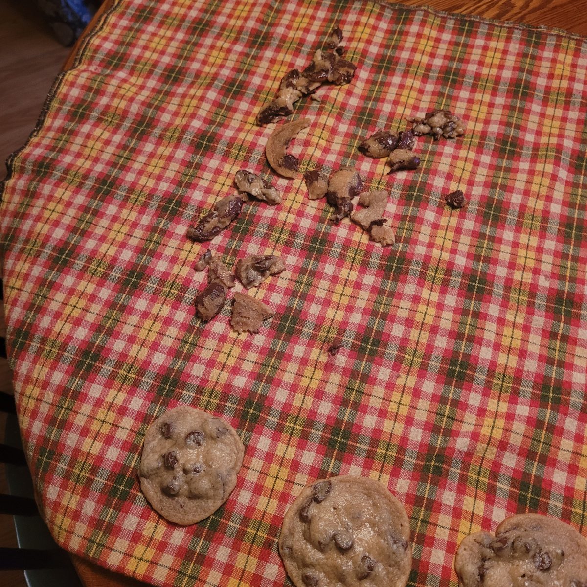 Chocolate chip cookies and cookie crumbs are scattered on a red and yellow plaid cloth placed on a wooden surface.