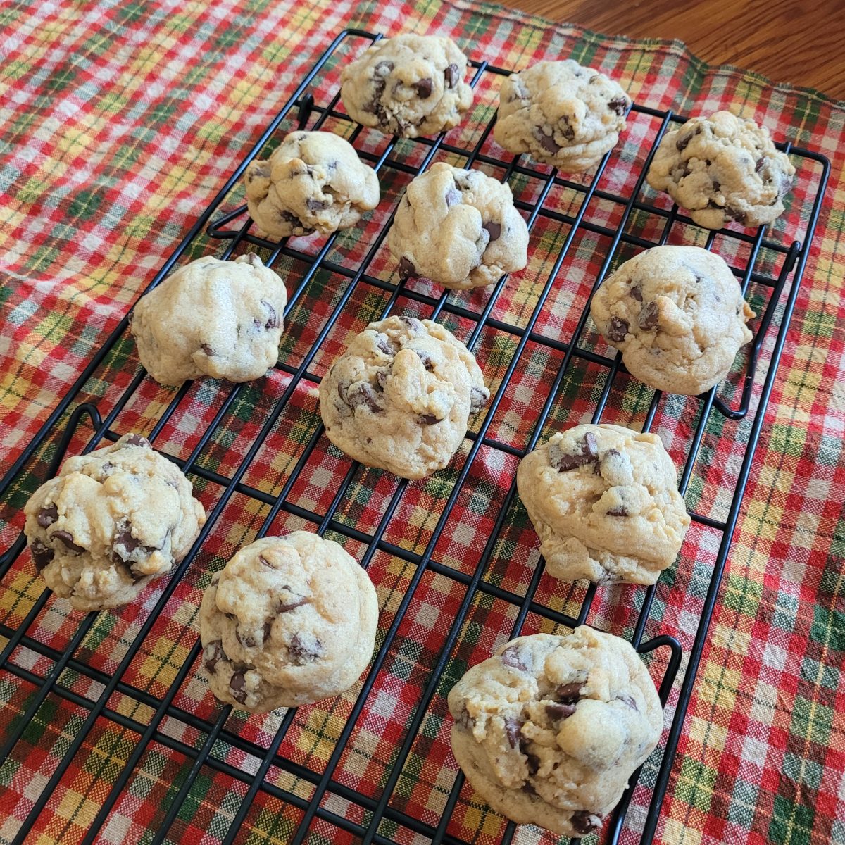 A cooling rack with freshly baked chocolate chip cookies sits on a red and green plaid tablecloth.