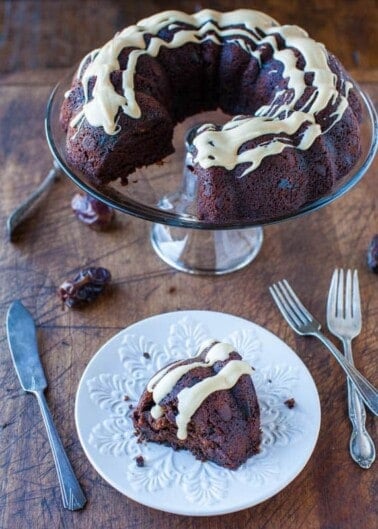 A chocolate bundt cake with white glaze on a glass stand, with a slice served on a plate beside two forks.