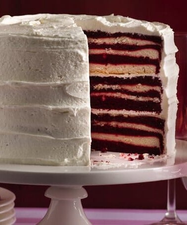 A multi-layered red velvet cake with white frosting on a cake stand, with wine glasses in the background.