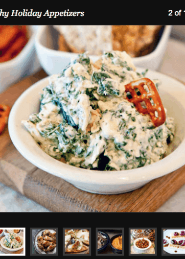 A bowl of creamy spinach dip with a cracker sticking out, surrounded by other assorted appetizers in the background.