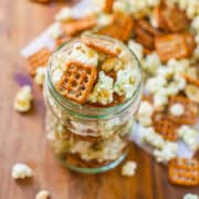 A jar filled with popcorn and pretzels on a wooden surface with scattered snacks around it.