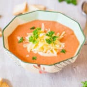 A bowl of creamy tomato soup garnished with shredded cheese and parsley, accompanied by pieces of bread on a linen cloth.