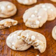 A batch of freshly baked white chocolate chip cookies on a wooden surface, one cookie broken open to show the melted chips inside.