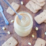 A jar of homemade peanut butter with a spoon in it, surrounded by biscuits and peanuts.