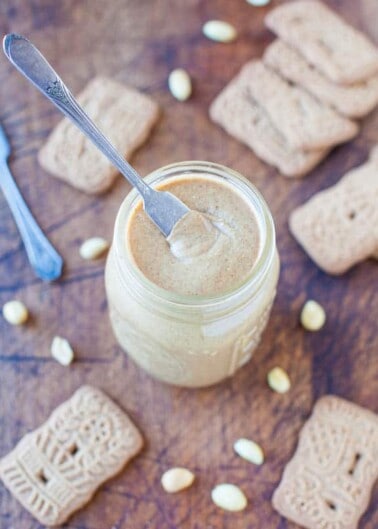 A jar of homemade peanut butter with a spoon in it, surrounded by biscuits and peanuts.