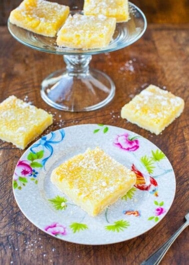 Lemon bars dusted with powdered sugar on a decorative plate with more in the background on a glass stand.