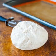 A ball of bread dough on a wooden surface with a baking sheet in the background.
