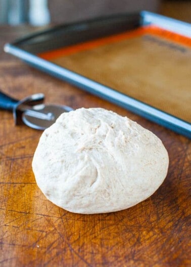 A ball of bread dough on a wooden surface with a baking sheet in the background.