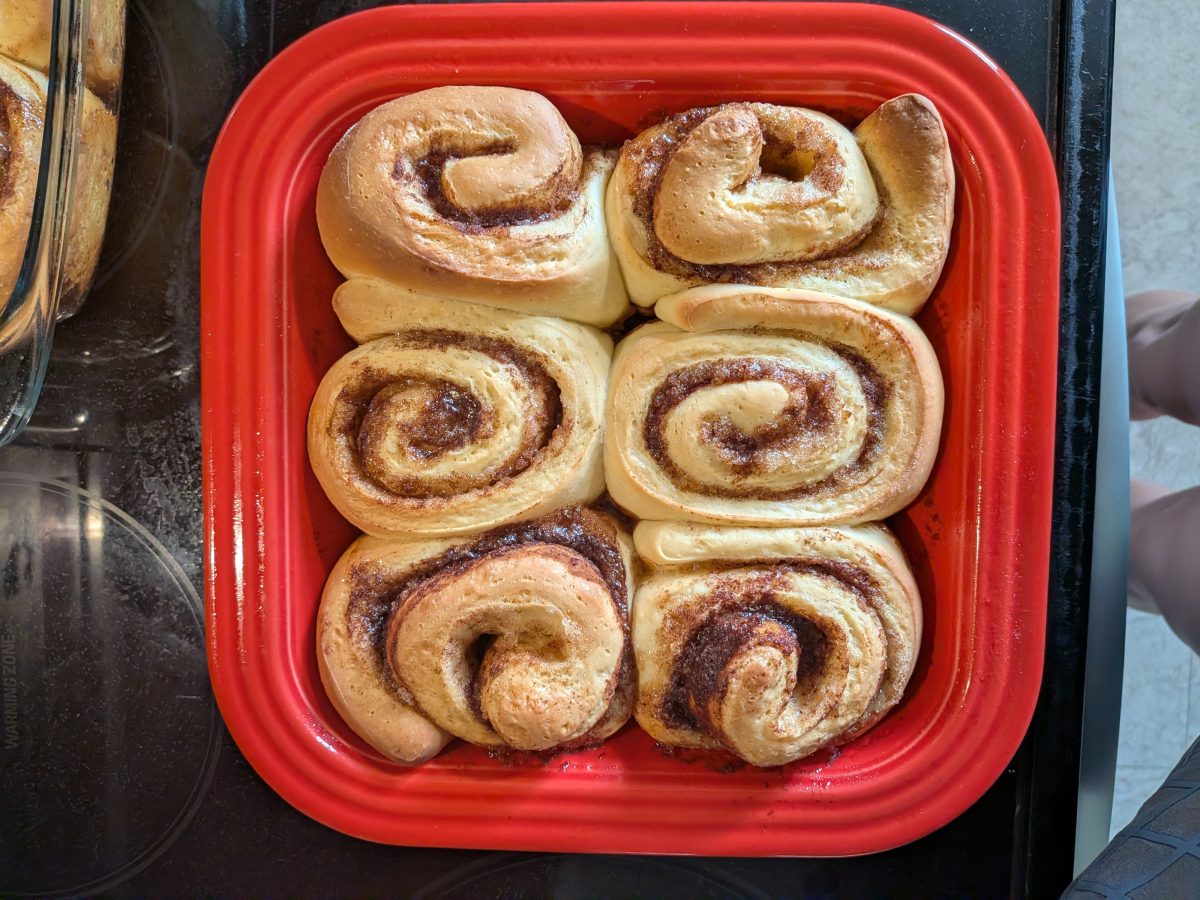 Six cinnamon rolls baked in a red square dish, viewed from above, sitting on a stovetop.