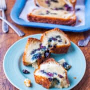 Sliced blueberry loaf cake served on a blue plate with forks and additional slices on a tray in the background.