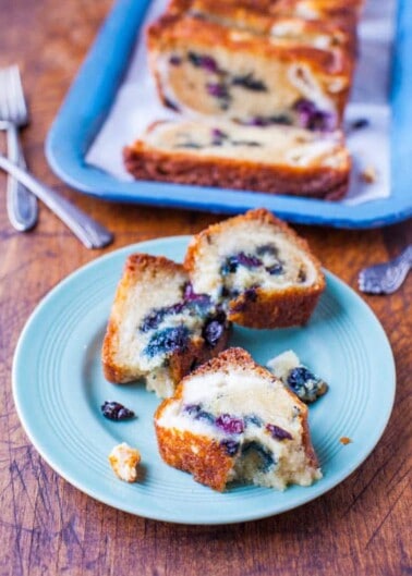 Sliced blueberry loaf cake served on a blue plate with forks and additional slices on a tray in the background.