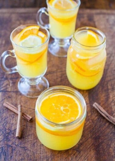 Three glasses of orange-colored drink garnished with citrus slices, accompanied by cinnamon sticks on a wooden surface.