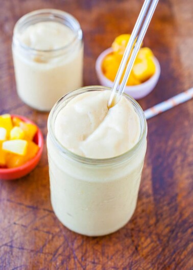 A mango smoothie in a glass jar with a metal straw, accompanied by mango chunks in the background.