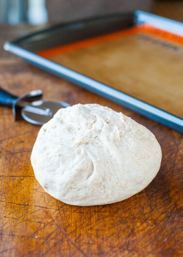A ball of raw bread dough on a wooden surface with a baking sheet and a pair of scissors in the background.