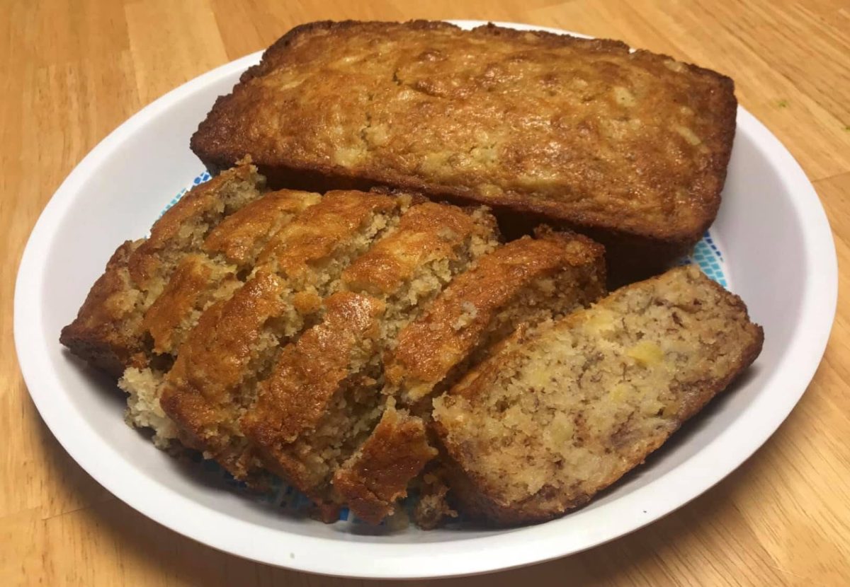 A white plate holds a loaf of banana bread, with several slices cut and arranged in front of the remaining unsliced portion.