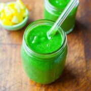 Two jars of green smoothie with a straw in one on a wooden surface, with a bowl of pineapple pieces in the background.