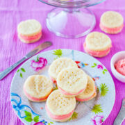 A plate of pink-filled sandwich cookies on a floral plate with a glass stand in the background.