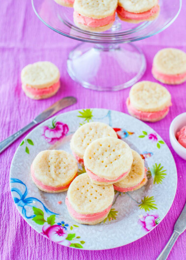 A plate of pink-filled sandwich cookies on a floral plate with a glass stand in the background.