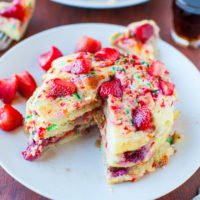 A slice of strawberry-studded cake with sprinkles on a white plate, accompanied by a fork and a small cup of syrup.