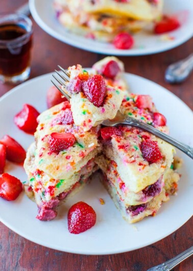 A plate of colorful, strawberry-topped funfetti pancakes with a fork and a small cup of syrup in the background.