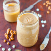 Two jars of homemade peanut butter with scattered peanuts and a vintage spoon on a wooden table.
