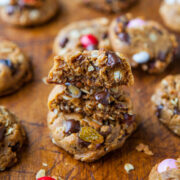 A variety of freshly baked cookies with chocolate chunks and nuts, one broken in half to reveal the chewy interior, on a wooden surface.