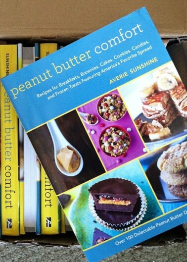 A cookbook titled "peanut butter comfort" by averie sunshine lies atop other books in a cardboard box.