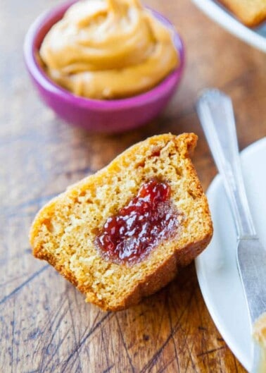 A slice of cornbread with a dollop of jelly on a wooden surface next to a knife and a small bowl of spread.