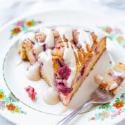 A slice of raspberry cake with icing drizzled on top, served on a floral plate with a spoon on the side.