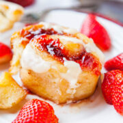 A freshly baked cinnamon roll with cream cheese frosting and strawberry toppings, accompanied by fresh strawberries on a white plate.