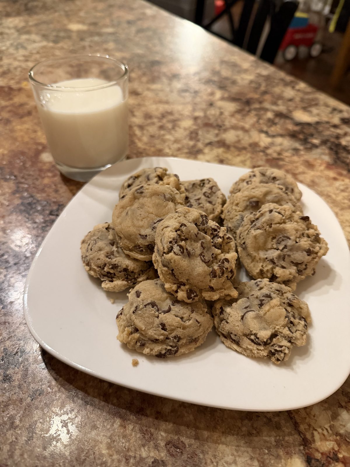 A white plate with several chocolate chip cookies sits on a marbled countertop next to a glass of milk.
