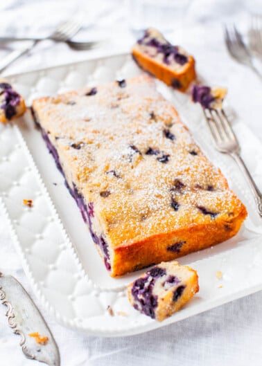 A blueberry loaf cake dusted with powdered sugar on a decorative plate with two forks.