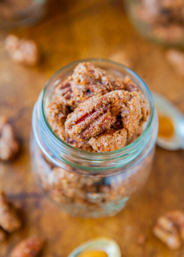 Candied pecans in a glass jar with more nuts and a honey dipper in the background.