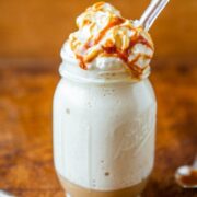 Vanilla milkshake in a mason jar, topped with whipped cream and caramel sauce, accompanied by a spoon with caramel on a wooden surface.