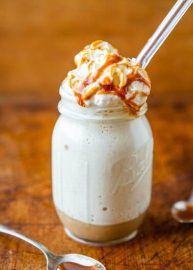 Vanilla milkshake in a mason jar, topped with whipped cream and caramel sauce, accompanied by a spoon with caramel on a wooden surface.