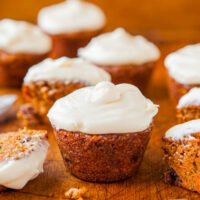 A selection of freshly baked carrot cupcakes topped with cream cheese frosting, with one partially eaten on a wooden surface.
