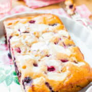 A freshly baked blueberry loaf cake with a sugar glaze, presented on a decorative plate with a pink and white checked cloth beneath it.