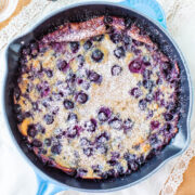 A freshly baked blueberry clafoutis in a cast iron skillet dusted with powdered sugar, served on a lace tablecloth.