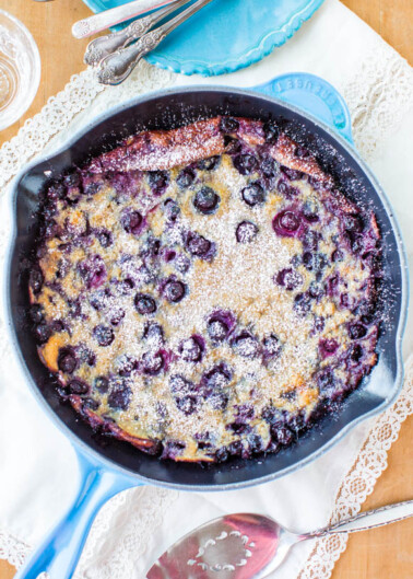 A freshly baked blueberry clafoutis in a cast iron skillet dusted with powdered sugar, served on a lace tablecloth.