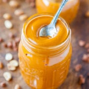 A jar of creamy peanut butter with a spoon inserted, on a wooden surface with peanuts scattered around.
