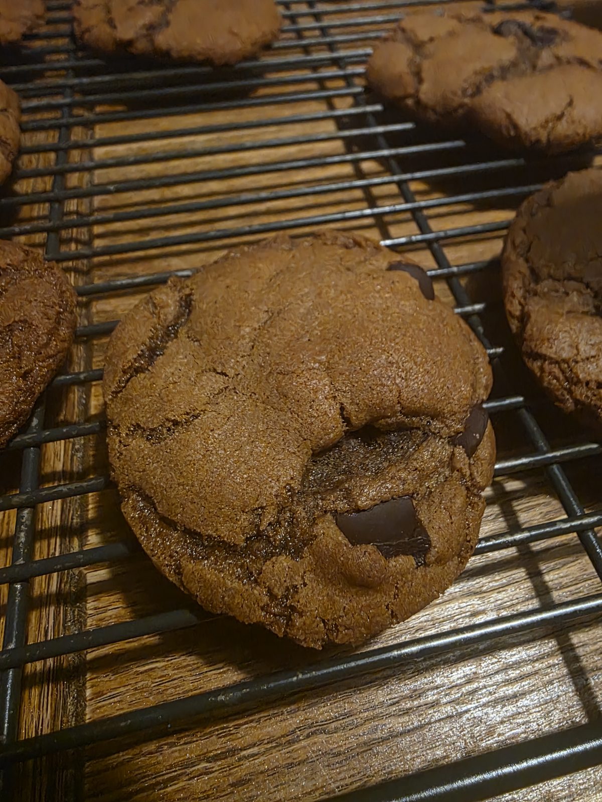A close-up of a chocolate cookie with visible cracks and chunks, cooling on a black wire rack on a wooden surface.