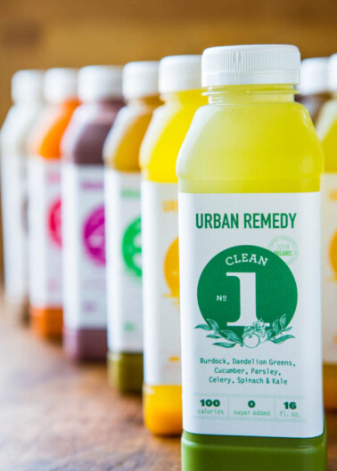 A line of colorful juice bottles with the nearest one labeled "urban remedy clean" with ingredients like cucumber, parsley, and kale listed.