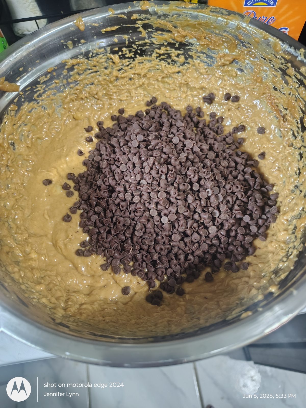 A large metal bowl containing cookie dough with a mound of chocolate chips on top, ready to be mixed.
